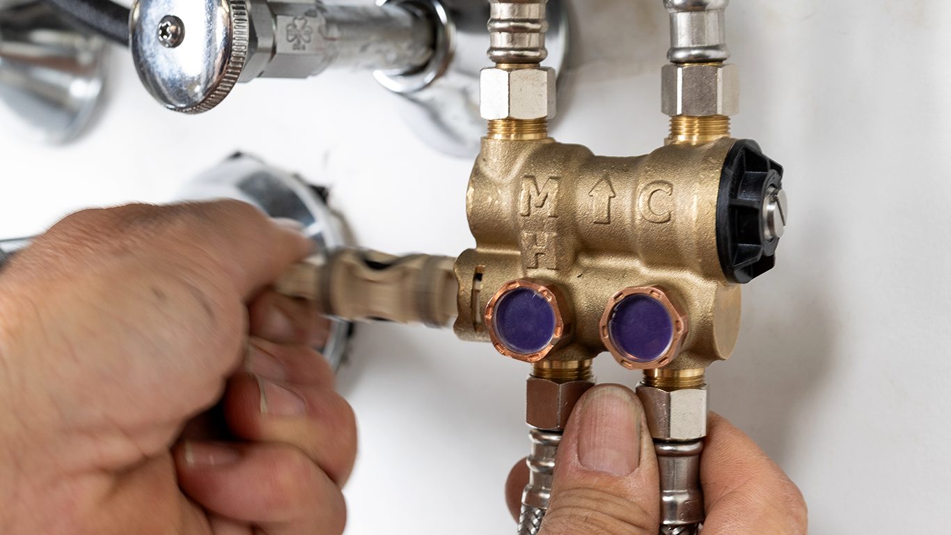 Close-up of hands working on a brass plumbing manifold with various pipes and valves.