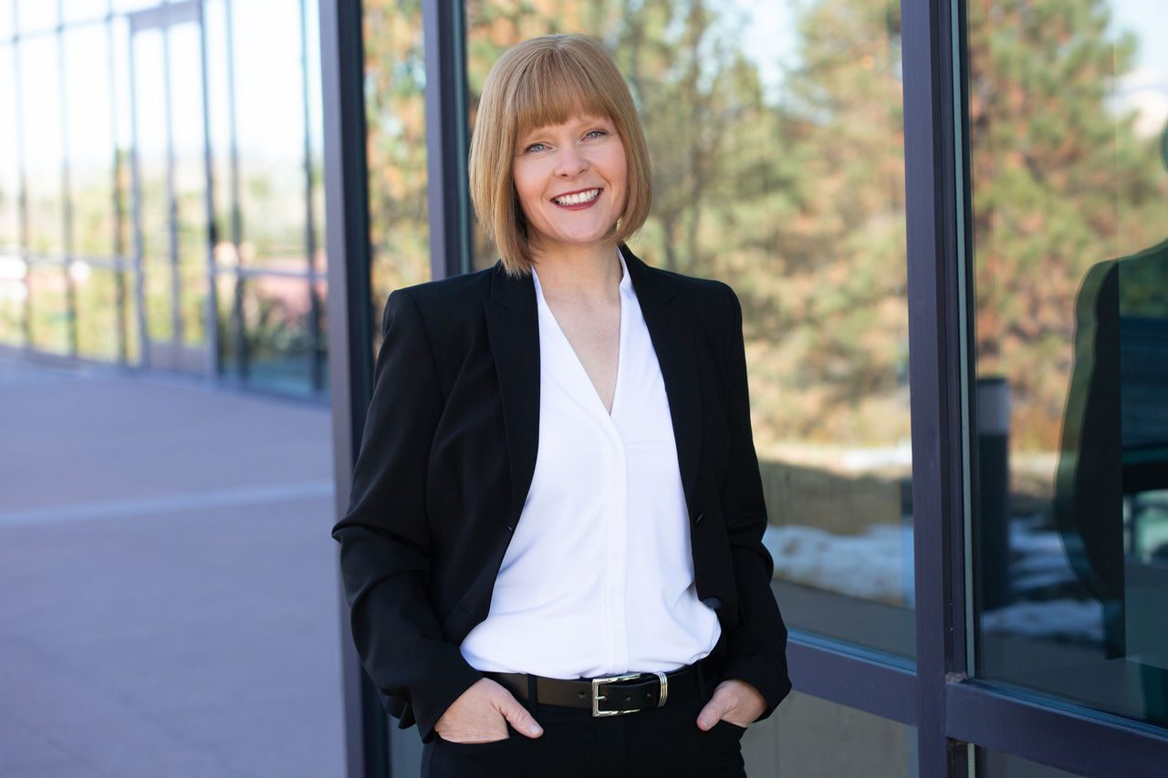 A smiling woman in a black blazer and white shirt stands in front of a modern glass building.