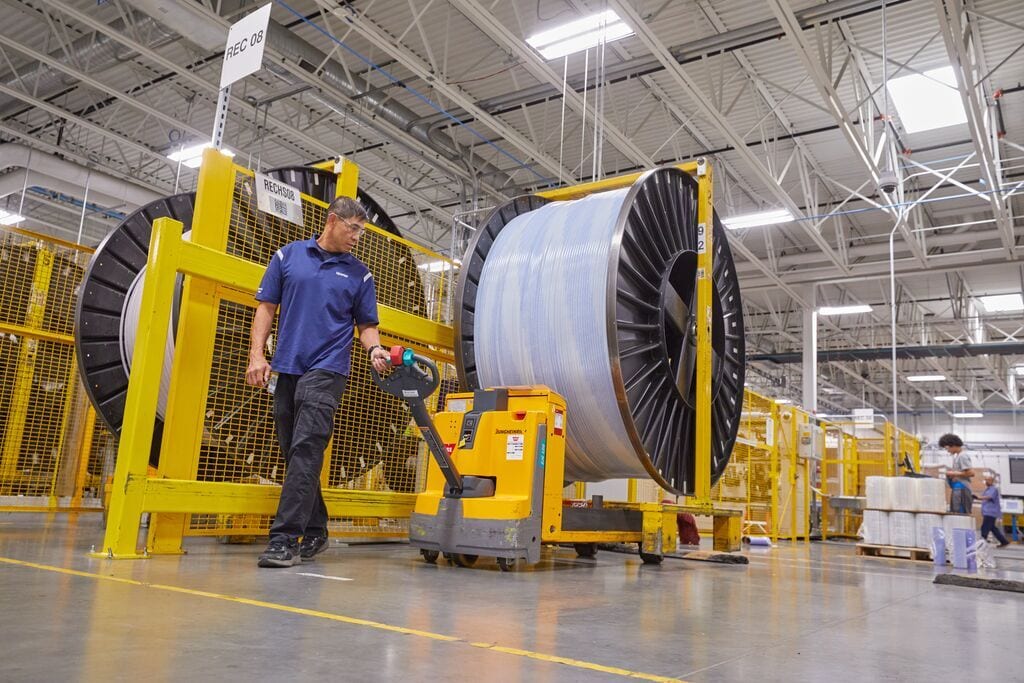 Man in a factory operating an electric pallet jack to move a large industrial cable spool.