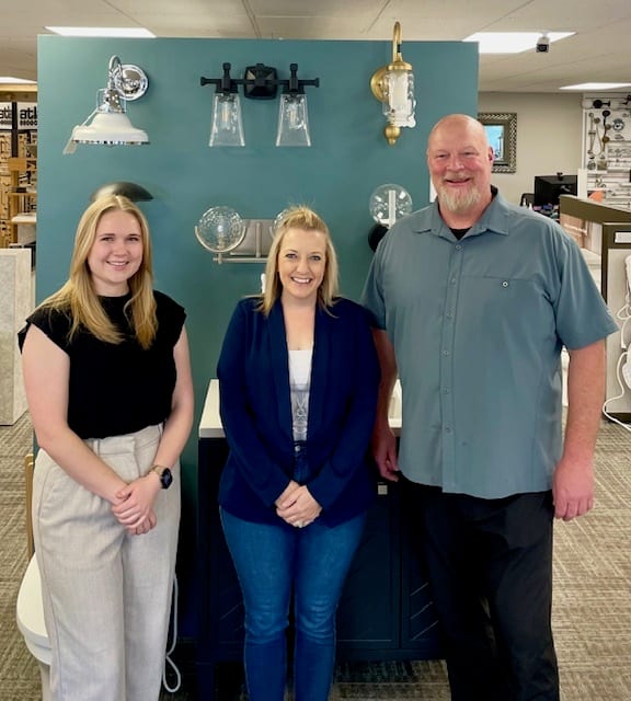 Two women and a man smile in a showroom featuring light fixtures and a vanity.