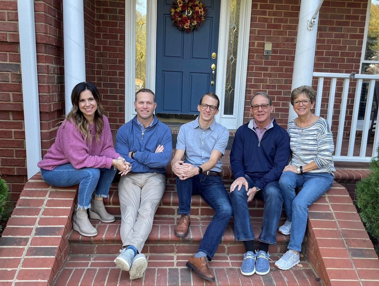 Five smiling people sitting on brick steps in front of a brick house with a blue door.