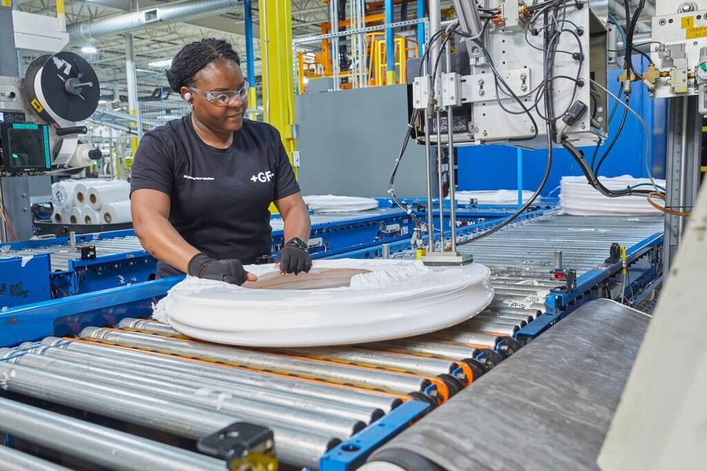 Woman in a factory working on a conveyor belt, handling a large white circular product, wearing safety glasses.