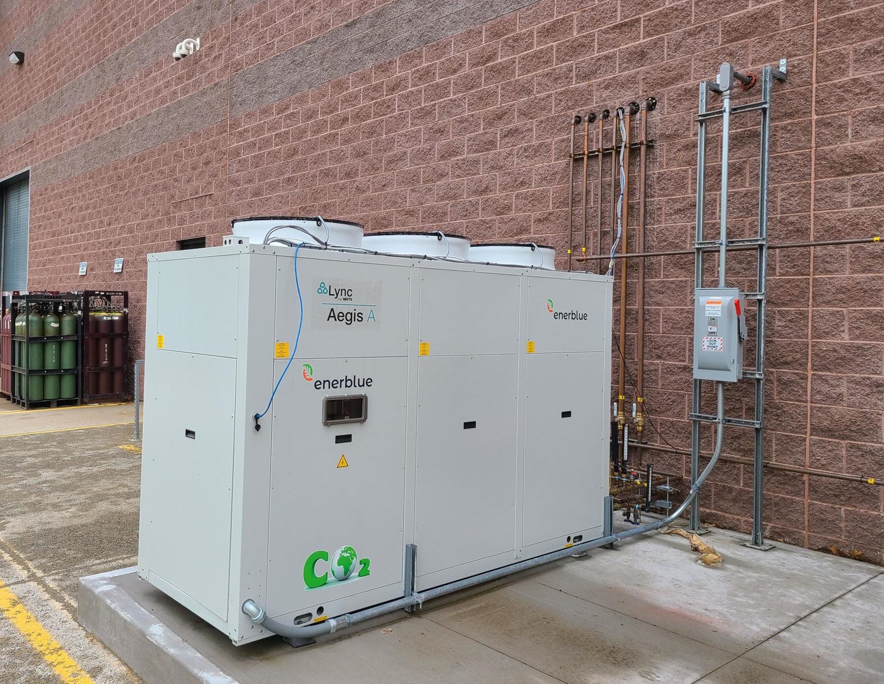 Large Lync Aegis A / Enerblue CO2 HVAC unit outdoors by a brick building with pipes.