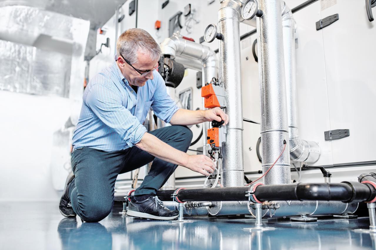Man in industrial setting kneels to adjust controls on large silver pipes.