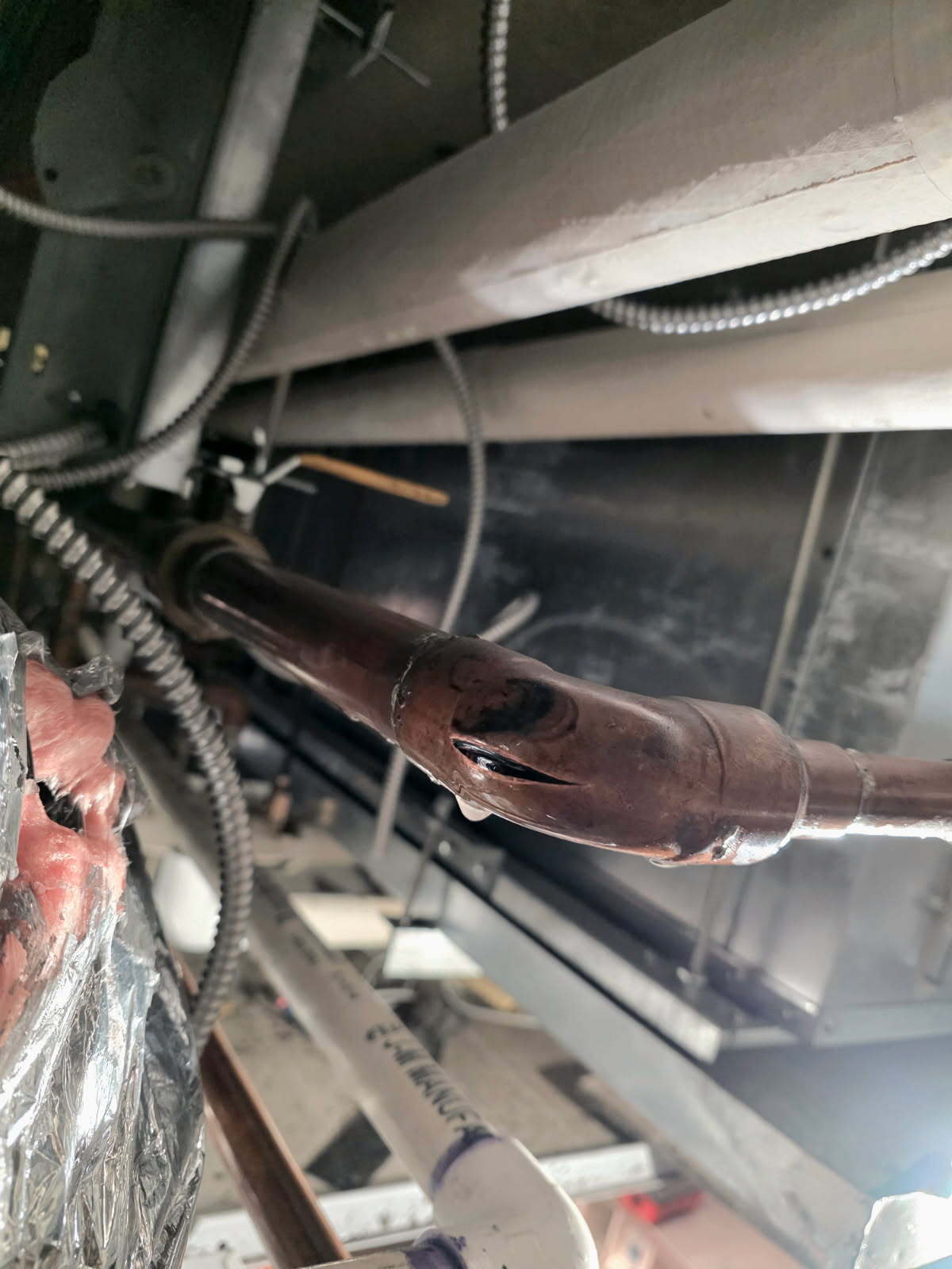 A burst copper pipe with water leaking from a large slit, surrounded by other pipes and ceiling elements.