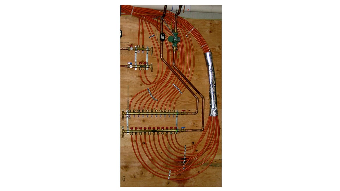 Radiant floor heating manifold system with orange PEX tubing, copper pipes, and pump on wood.