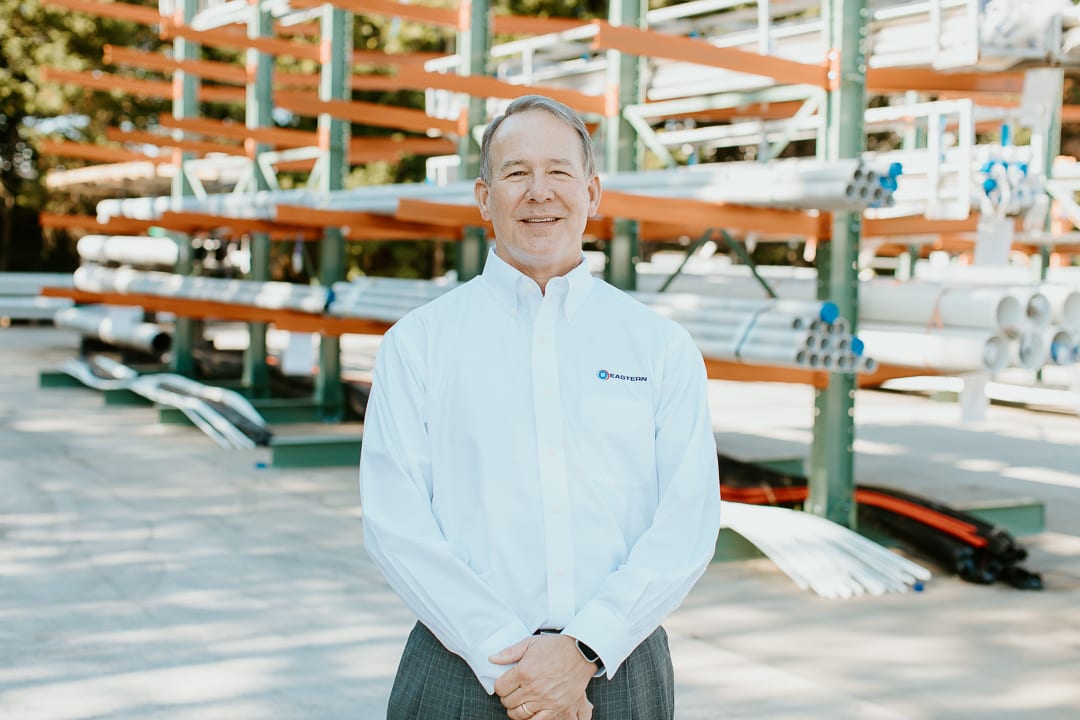Smiling man in white 'EASTERN' shirt stands before industrial pipe shelving.