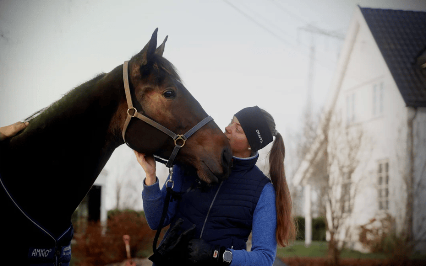 A woman in a beanie and vest kisses a dark horse in a halter, with a white house behind.