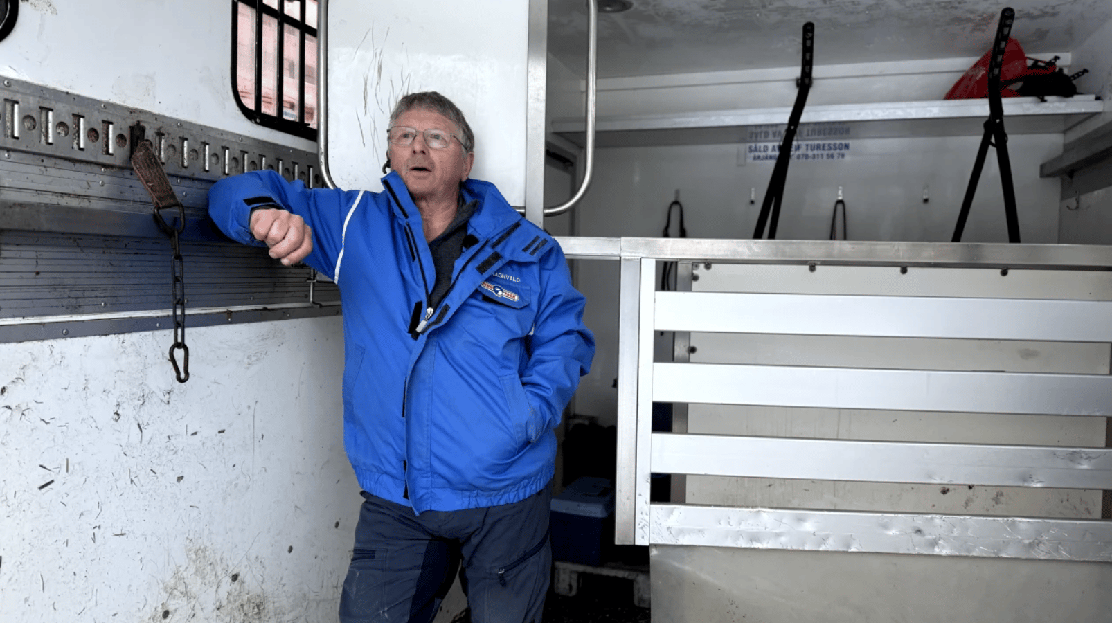 An older man in a blue jacket stands inside a utilitarian trailer, looking up and to the left.