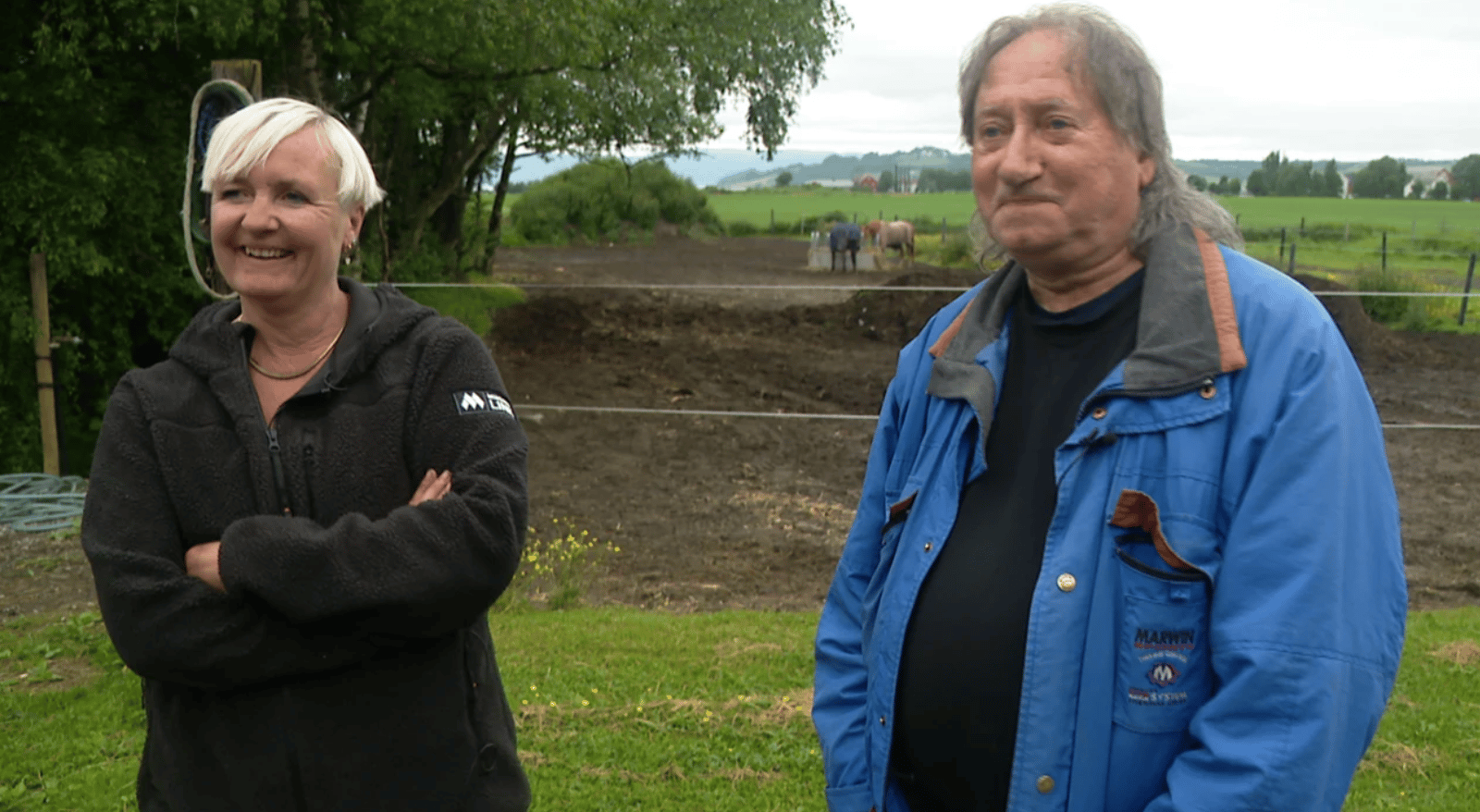 A smiling woman and a man stand outdoors on a farm with horses in the background.
