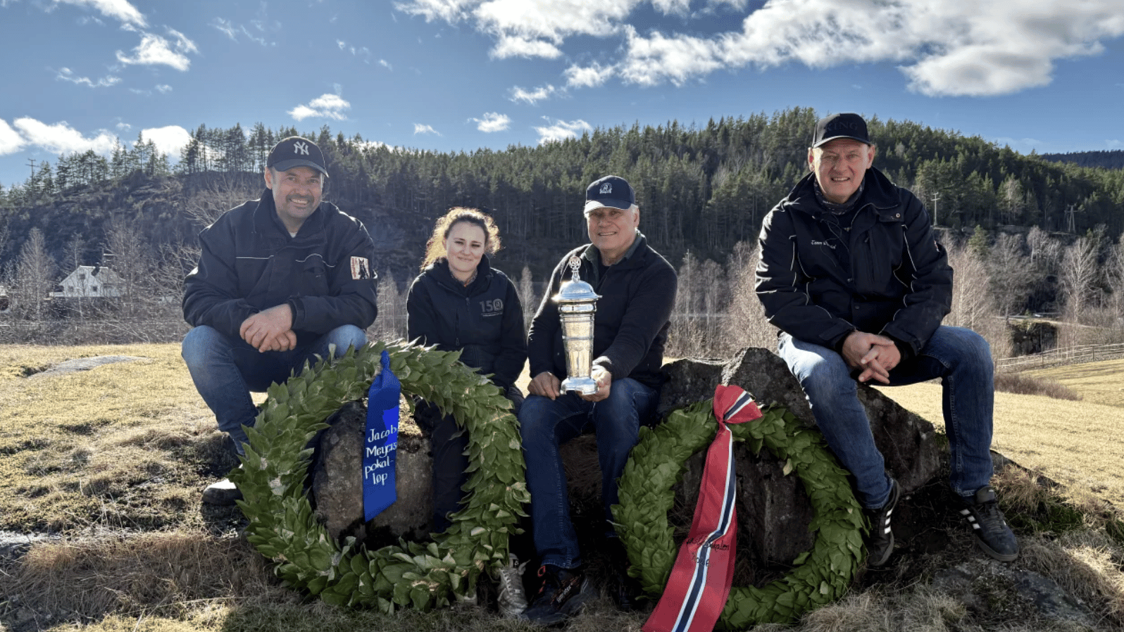 Four people, including a woman and three men, pose outdoors with a silver trophy and two laurel wreaths.