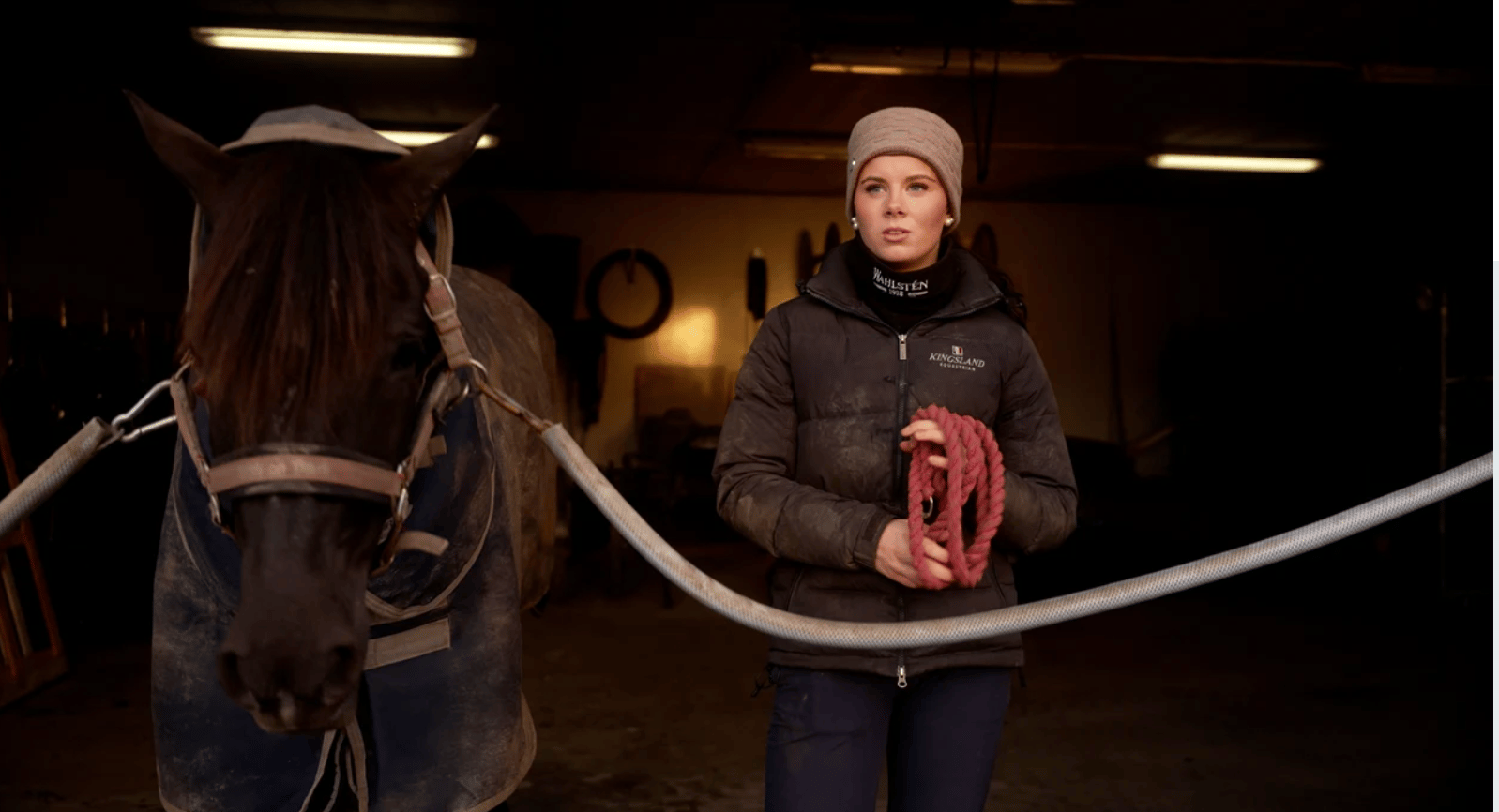 A young woman in a barn holds a red rope while standing next to a horse wearing a blanket.