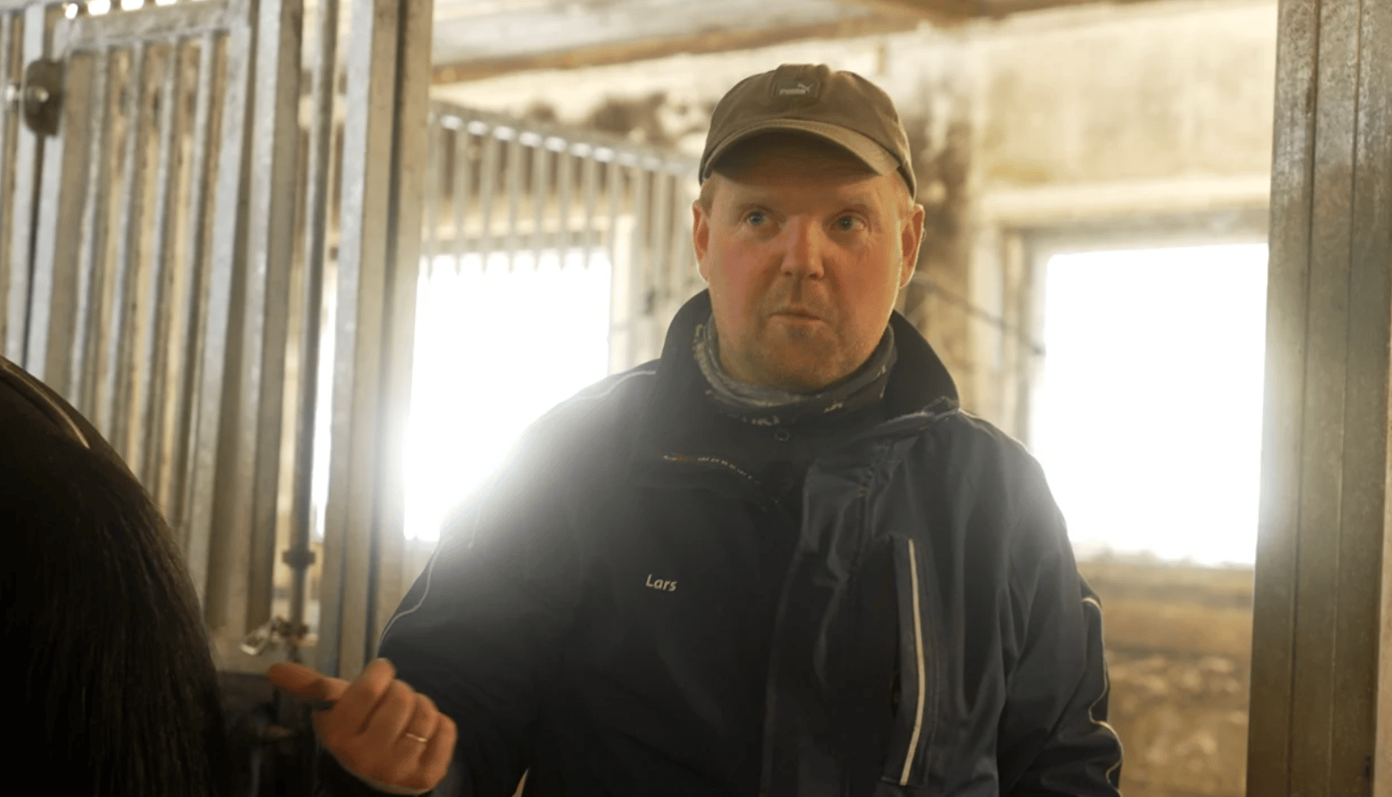 A man named Lars in a cap and jacket stands in a stable, looking ahead with a horse partly visible.