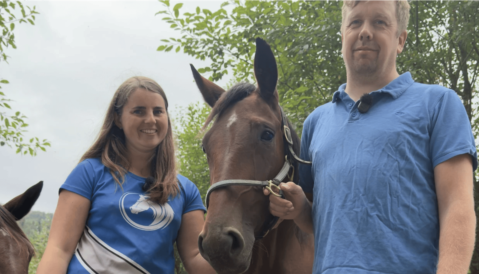 A woman, a man, and a horse standing outdoors, smiling, with trees in the background.
