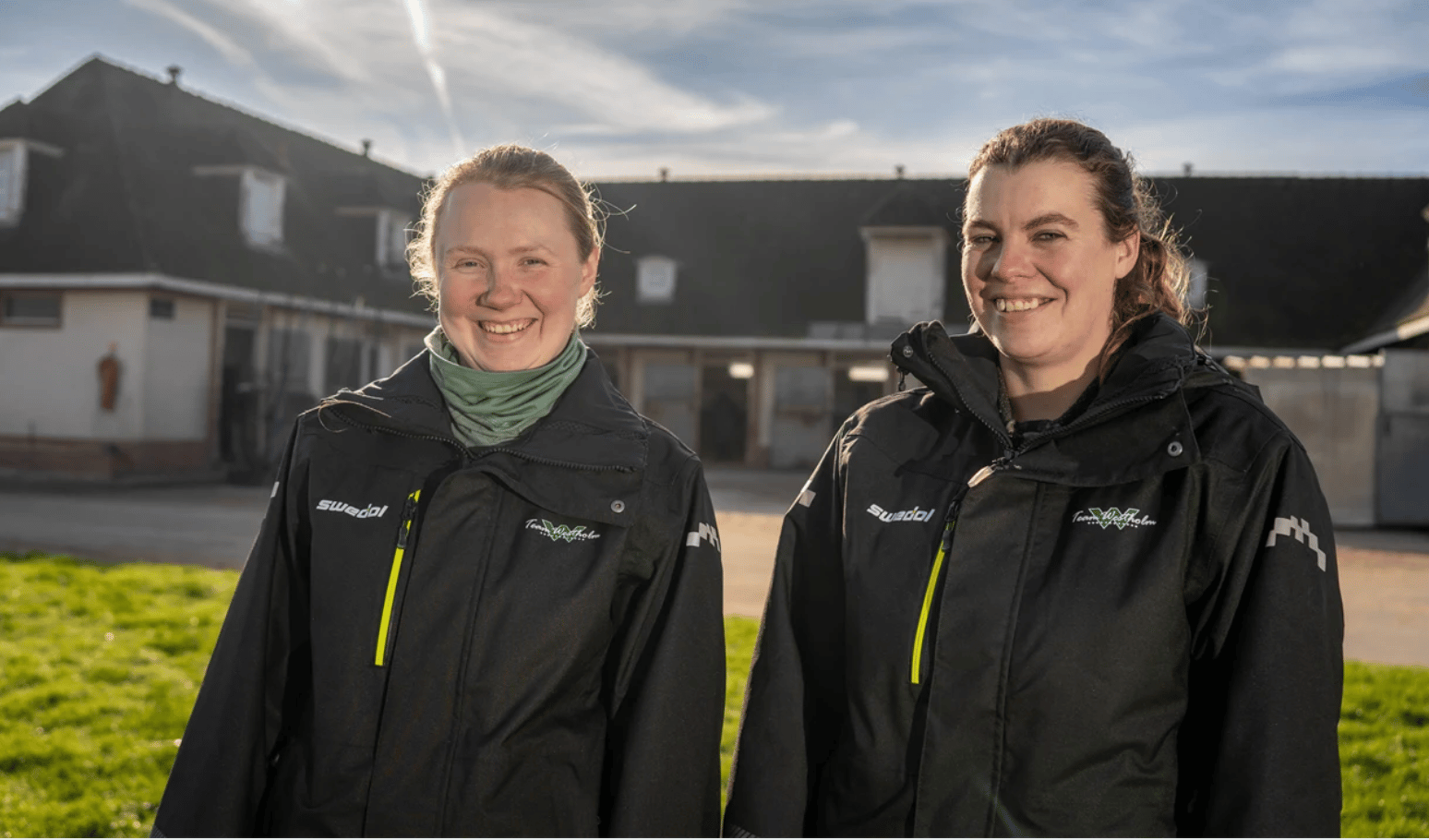 Two smiling women in black jackets stand outdoors in front of a farm building under a sunny sky.