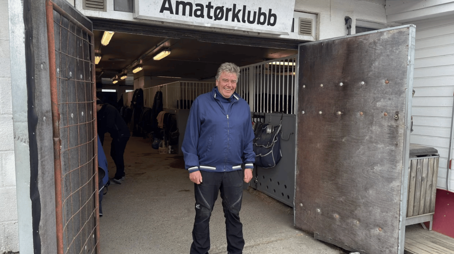 A smiling man in a blue jacket stands in a stable doorway with horse stalls visible in the background.