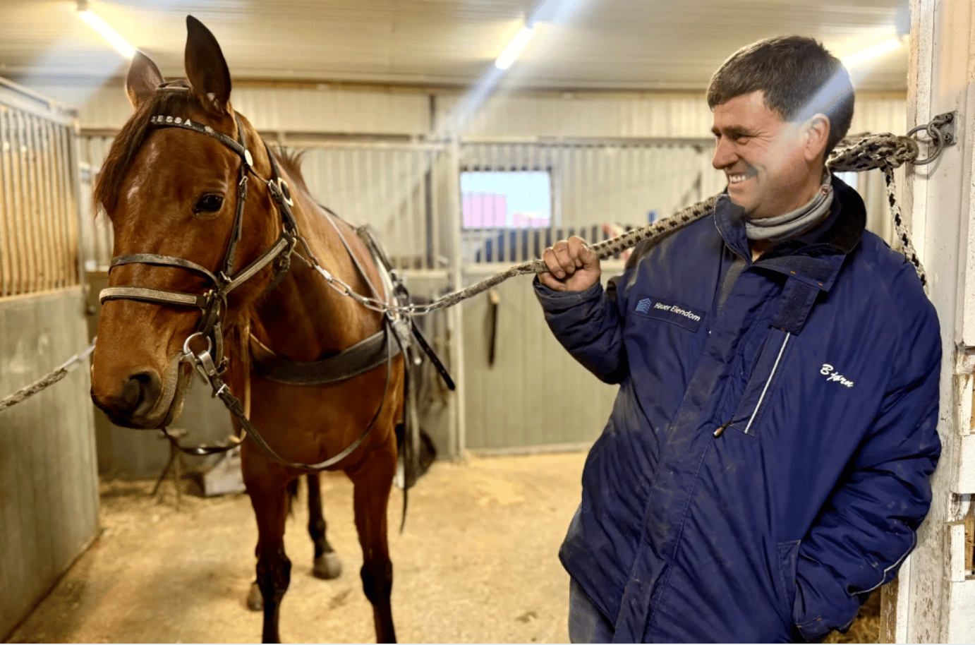 A smiling man in a blue jacket holds a rope connected to a brown horse's harness inside a stable.