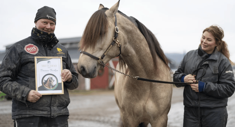 Man holding an "ÅRETS HEST Brenne Boken" award next to a dun horse and a woman.
