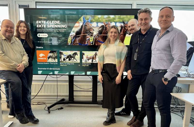 Six smiling colleagues stand in front of a large screen displaying horse racing content.