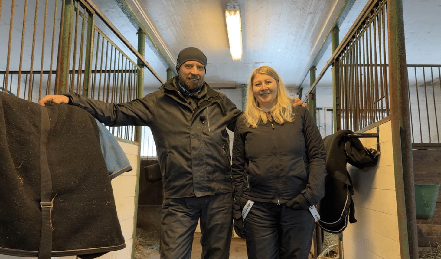 Two smiling people, a man and a woman, stand in a horse stable aisle between stalls.