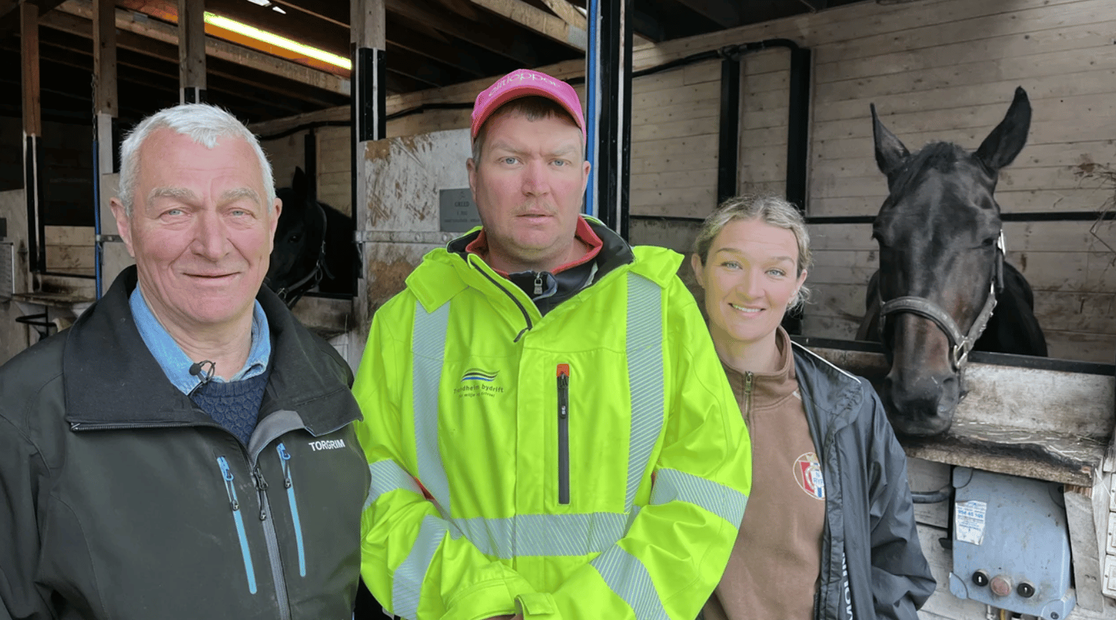 Three people, including a man in a bright yellow jacket, and a dark horse in a stable.