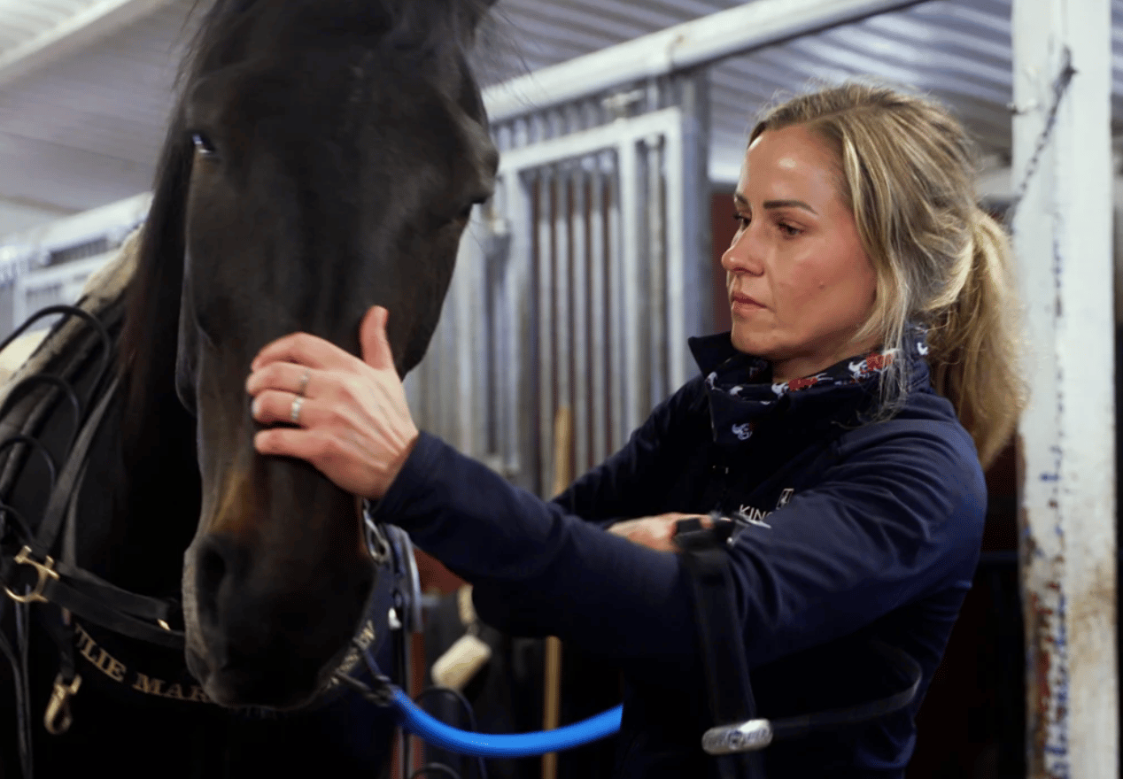 A woman gently strokes a dark horse's nose in a stable, showing a tender bond between them.