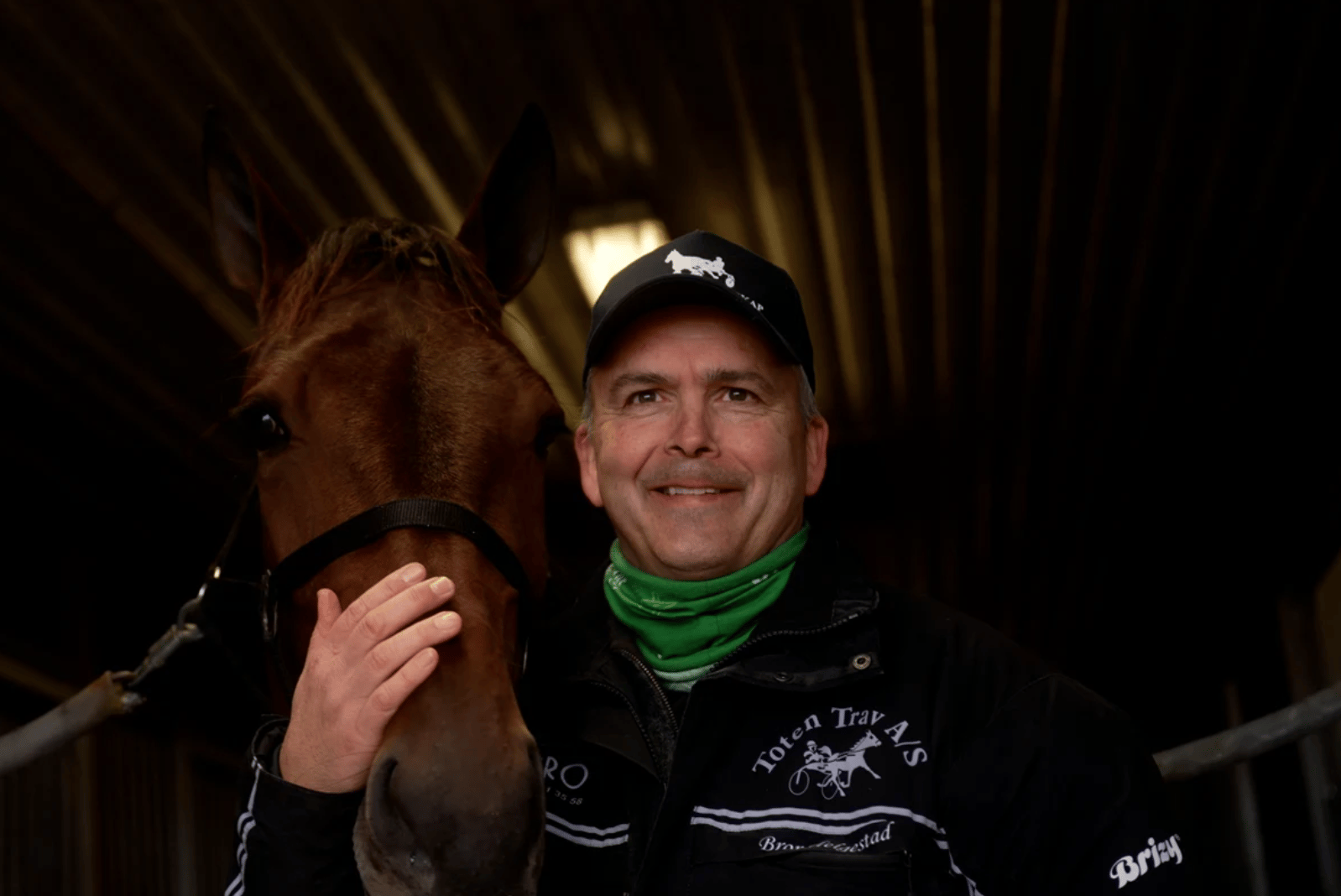A man smiling, wearing a cap and jacket, with his hand on a brown horse's face in a dimly lit stable.
