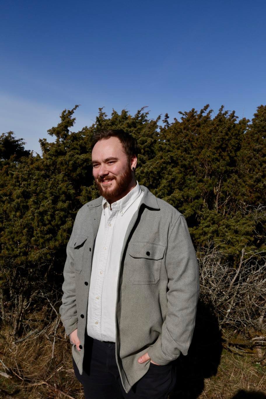 Smiling bearded man in gray jacket against trees and blue sky.