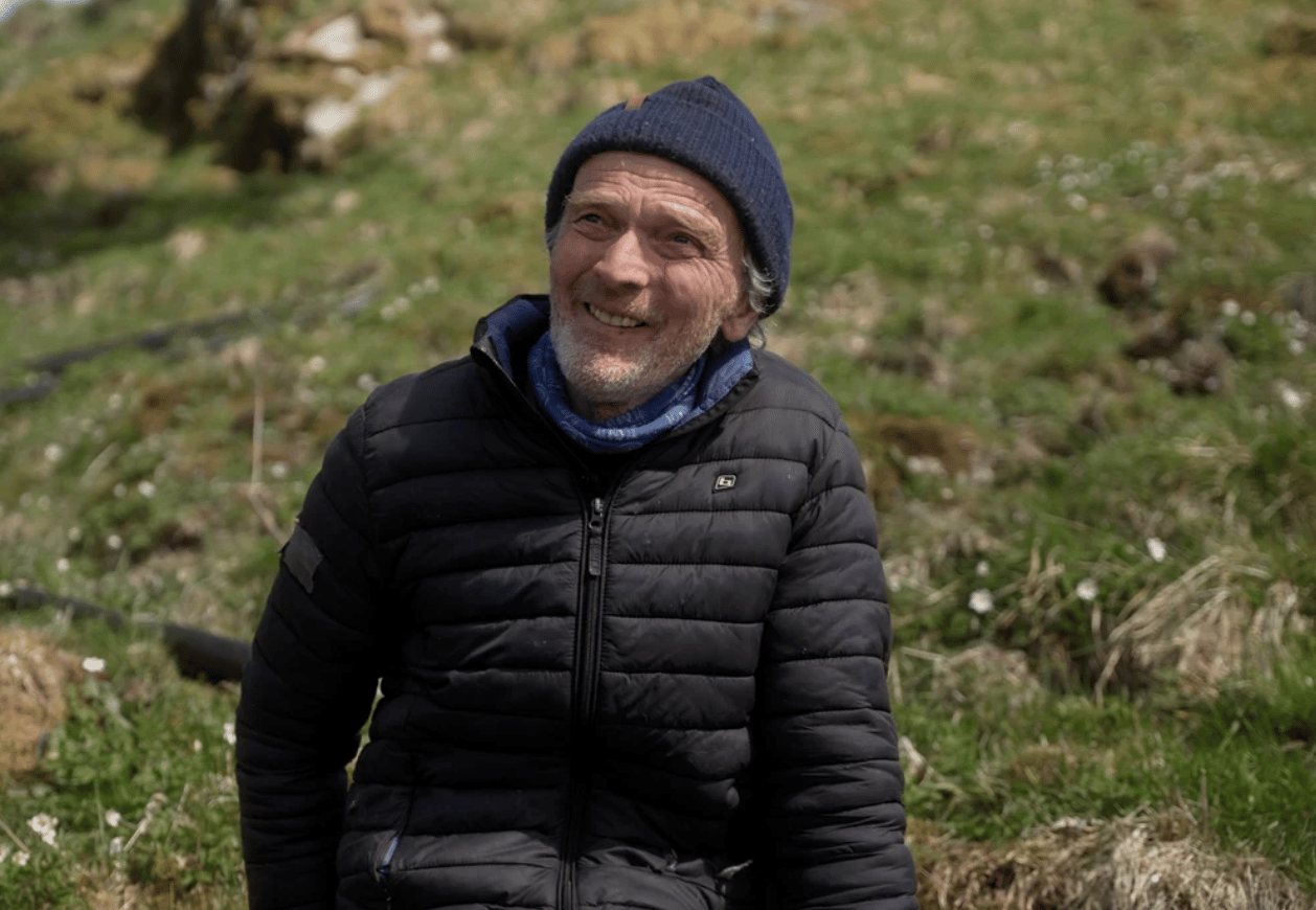 A smiling man in a blue beanie and black puffer jacket stands in a grassy field.