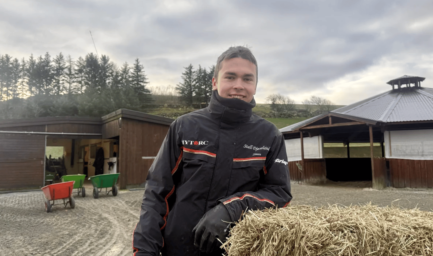 Smiling man in black jacket and gloves leaning on hay bale at a stable.