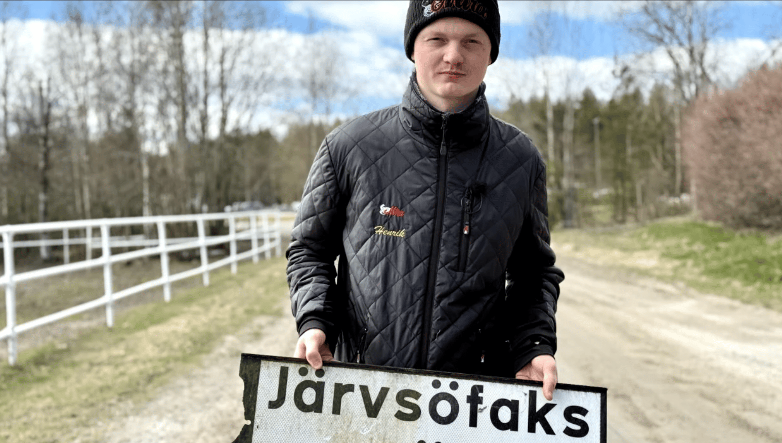 A young man in a black quilted jacket and beanie holds a sign that reads "Järvsöfaks" outdoors.