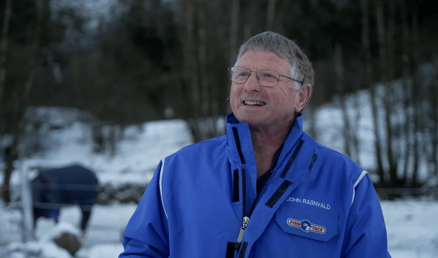 A smiling older man wearing glasses and a blue jacket stands outdoors in a snowy, wooded environment.
