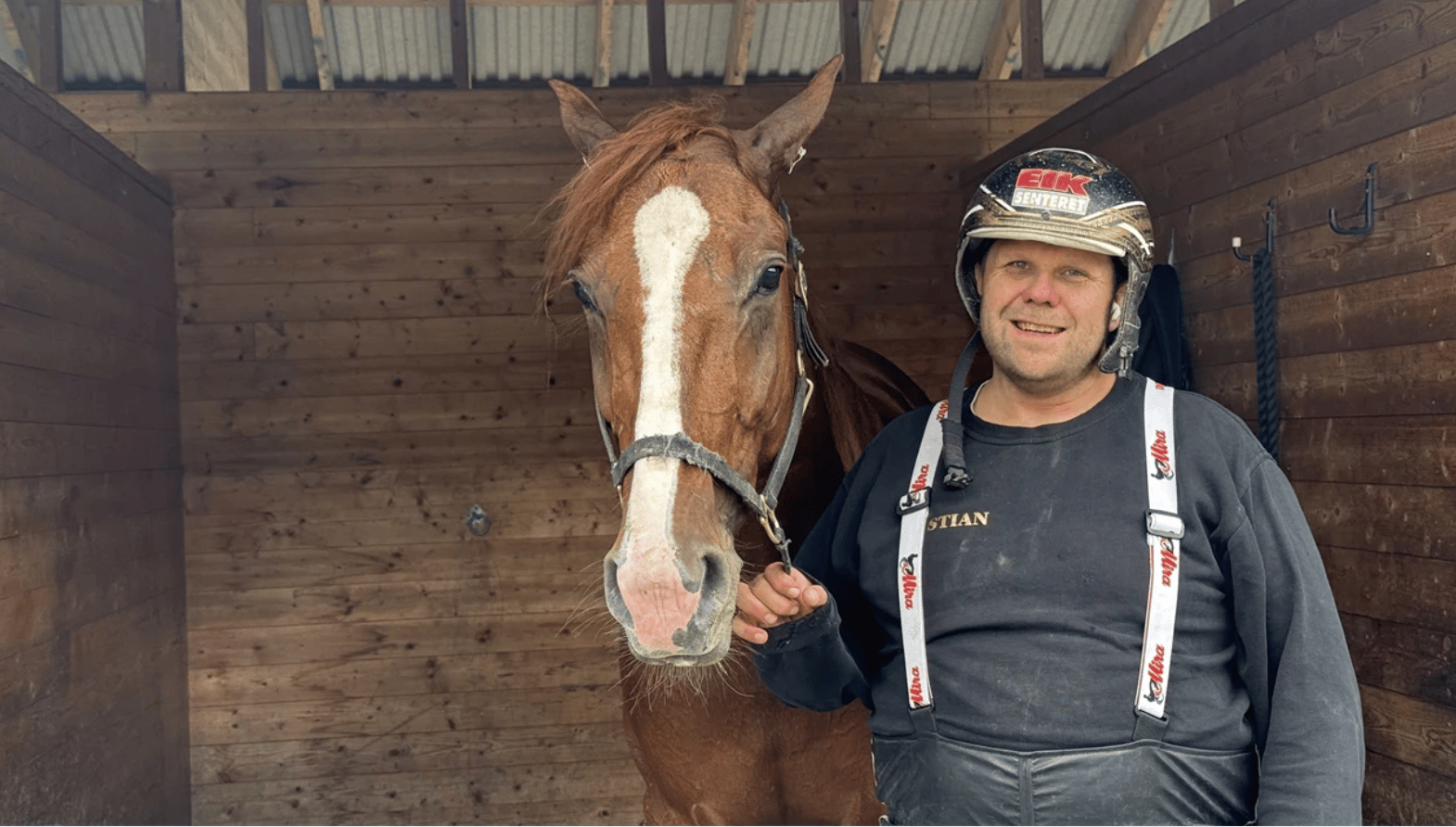 Man in a helmet and a brown horse with a white blaze inside a wooden stable.