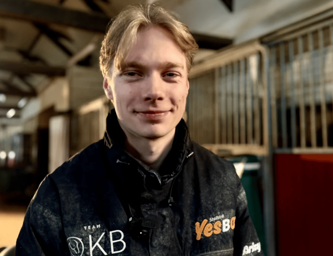 A smiling young man with blonde hair and a dark jacket stands in a stable or barn, looking forward.