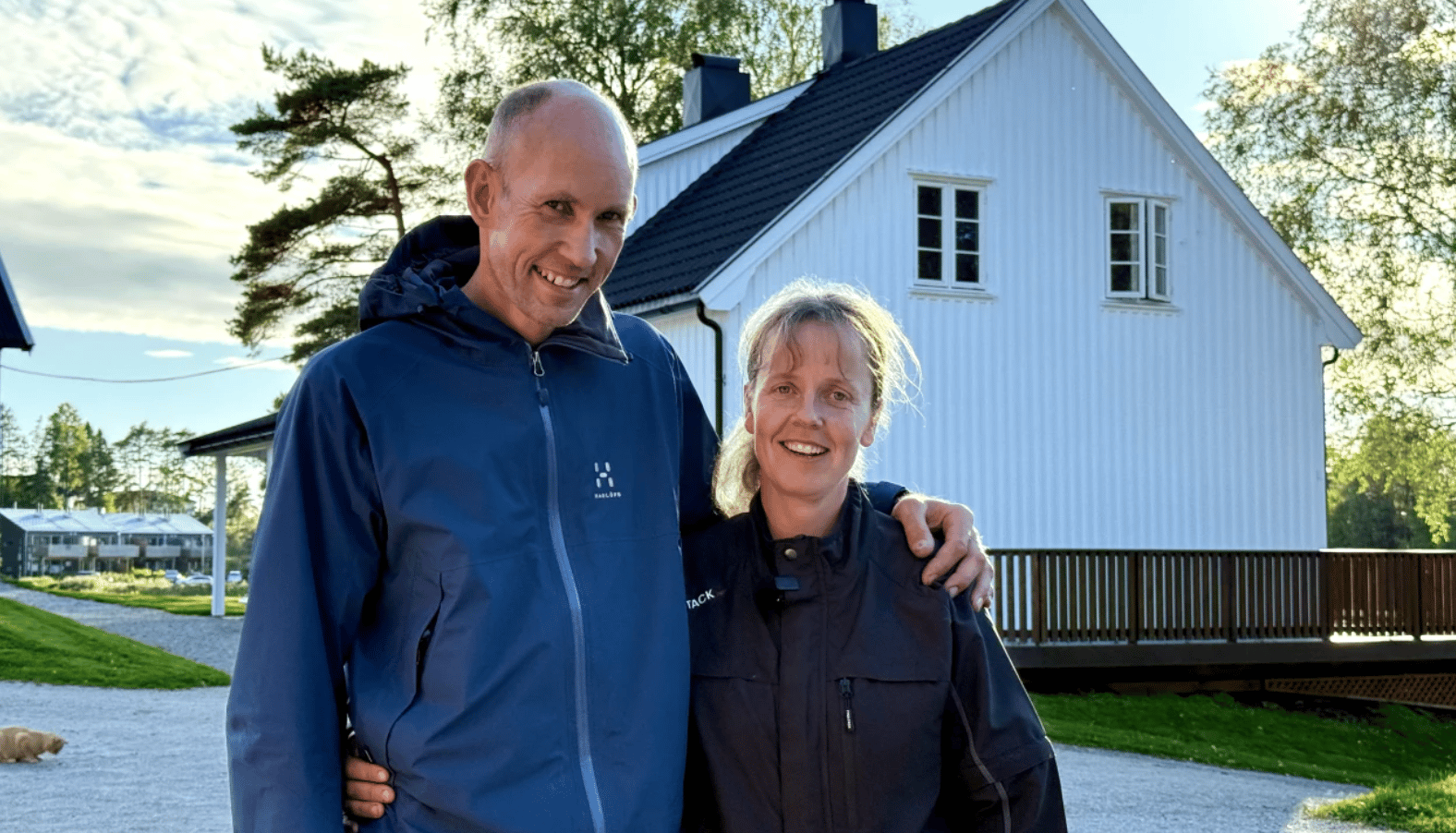 Man and woman smiling outdoors, standing in front of a white house with trees in the background.