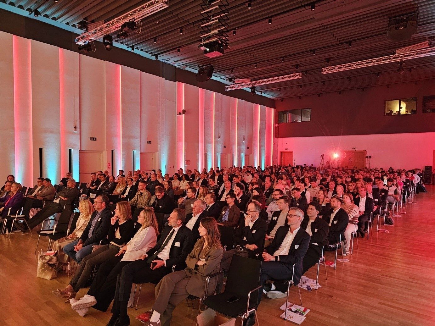 Large audience seated in rows at a conference or event, illuminated by colorful stage lighting.