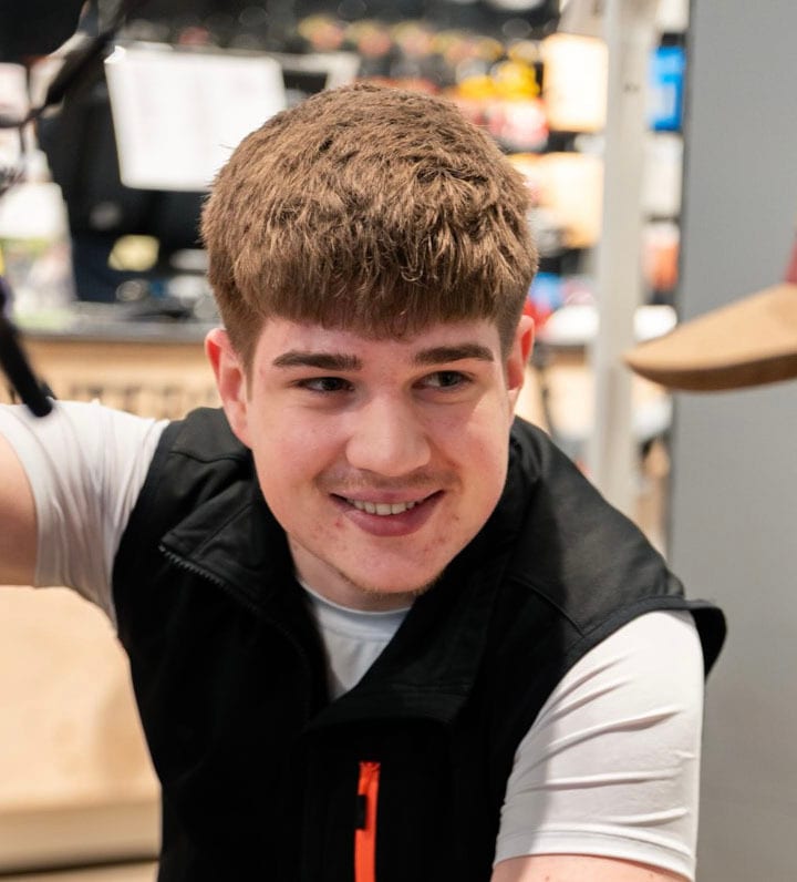 A young man with brown hair and a black vest smiles while looking to his right.