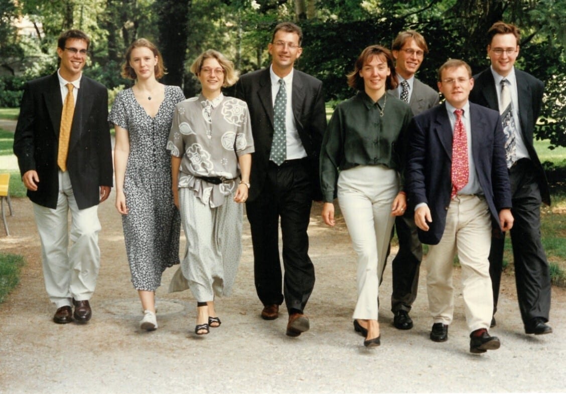 Eight individuals, four men in suits and four women in dresses/smart casual, walk together outdoors.