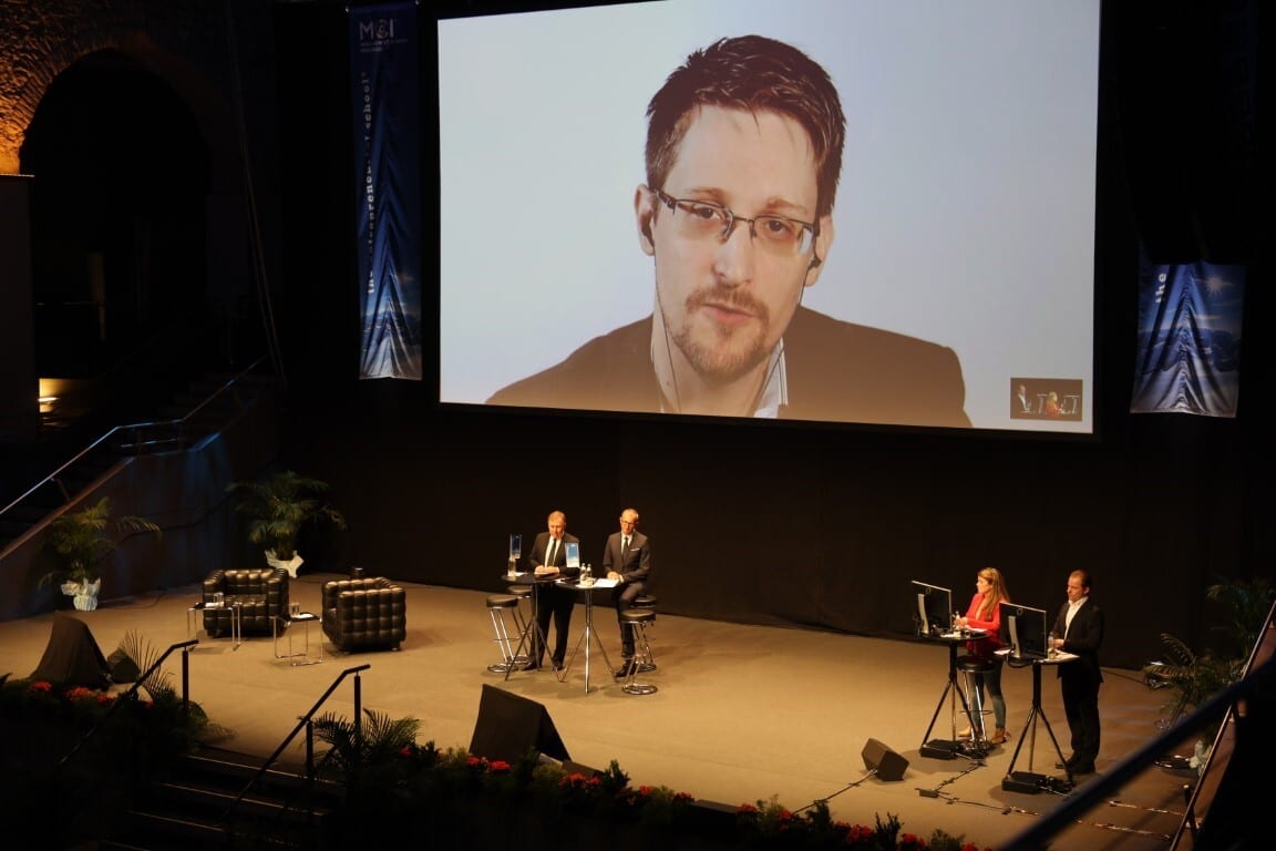 Edward Snowden appears on a large screen during a conference, with two men speaking on stage and two people at computers to the right.