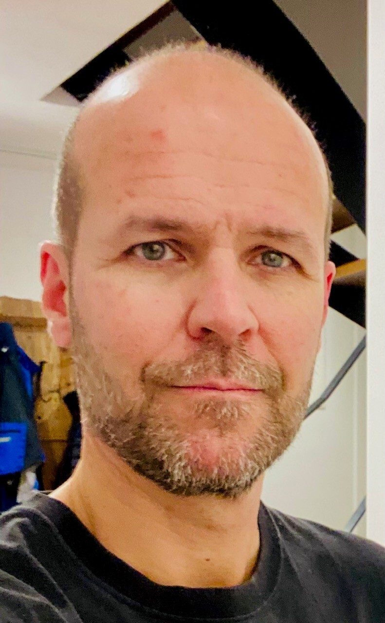 A close-up portrait of a bald man with blue-grey eyes and a light beard, looking directly at the camera.