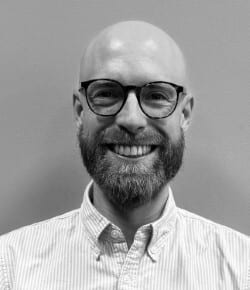Black and white headshot of a smiling bald man with glasses and a beard, wearing a striped shirt.