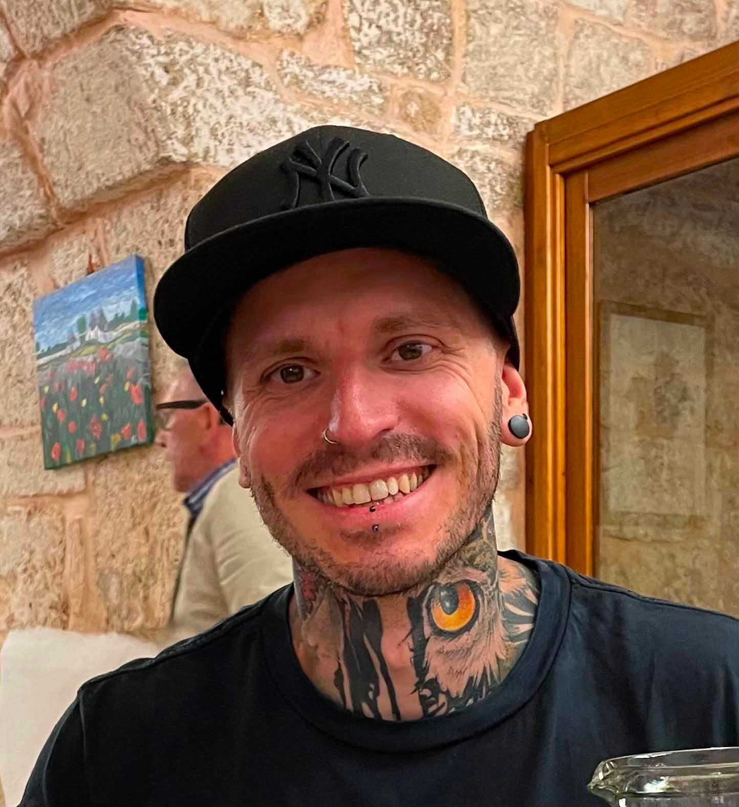 Smiling man in a black cap, with neck tattoos and piercings, against a stone wall background.