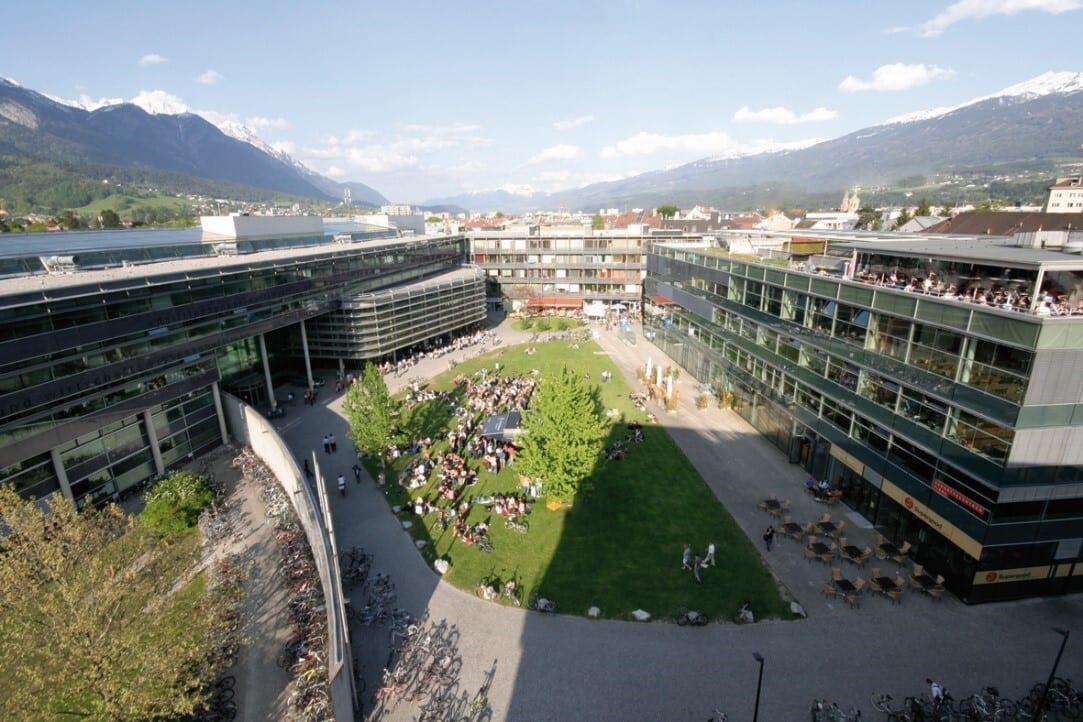 Aerial view of a modern university campus courtyard with people relaxing, surrounded by sleek buildings and mountains.