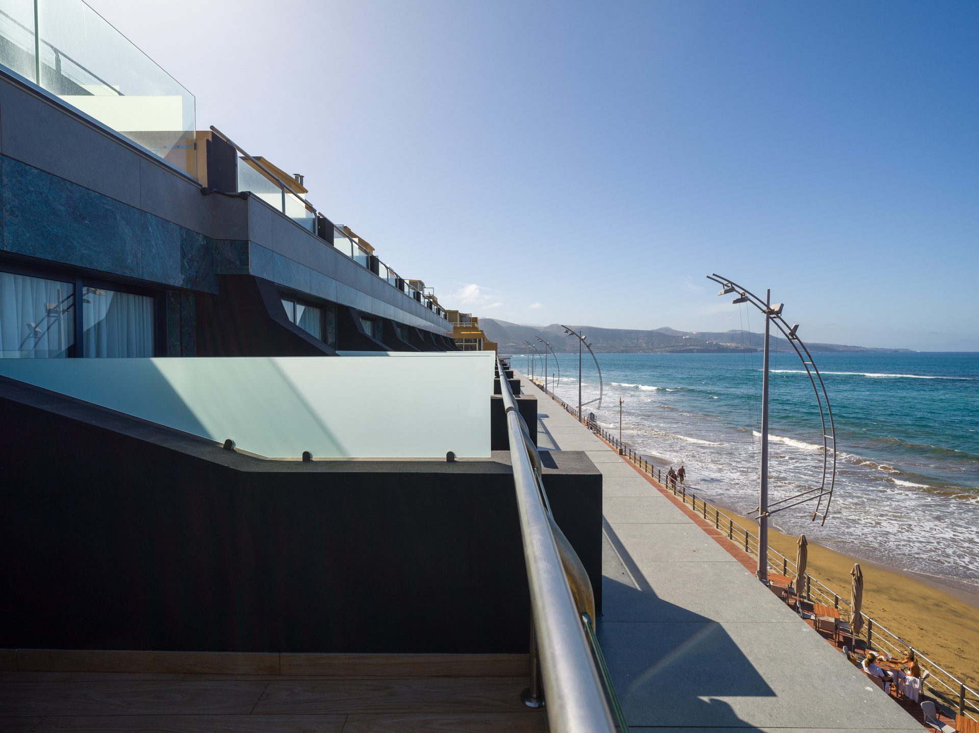 Modern hotel balconies overlook a sunny beach with ocean and mountains.