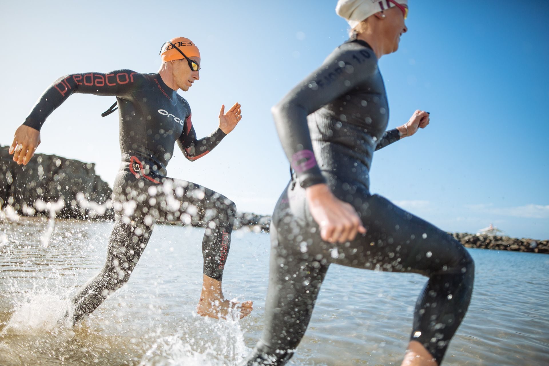 Man and woman in wetsuits running from water, splashing.