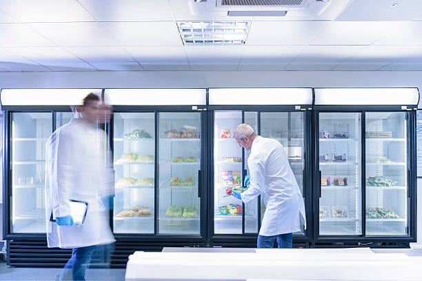 Two scientists in lab coats in a cold storage room with large fridges. One inspects a fridge, another walks past.