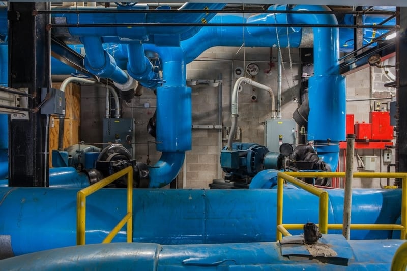 Industrial room with a complex system of large blue pipes, pumps, and machinery, featuring yellow safety railings.