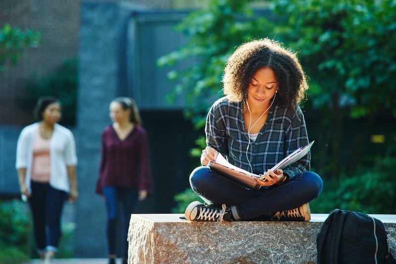 A young woman with curly hair sits on a stone bench, wearing headphones and studying outdoors.