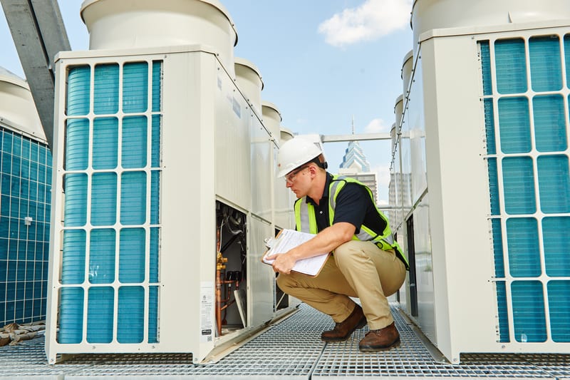 Technician in hard hat and safety vest inspecting an HVAC unit on a rooftop with city skyline.