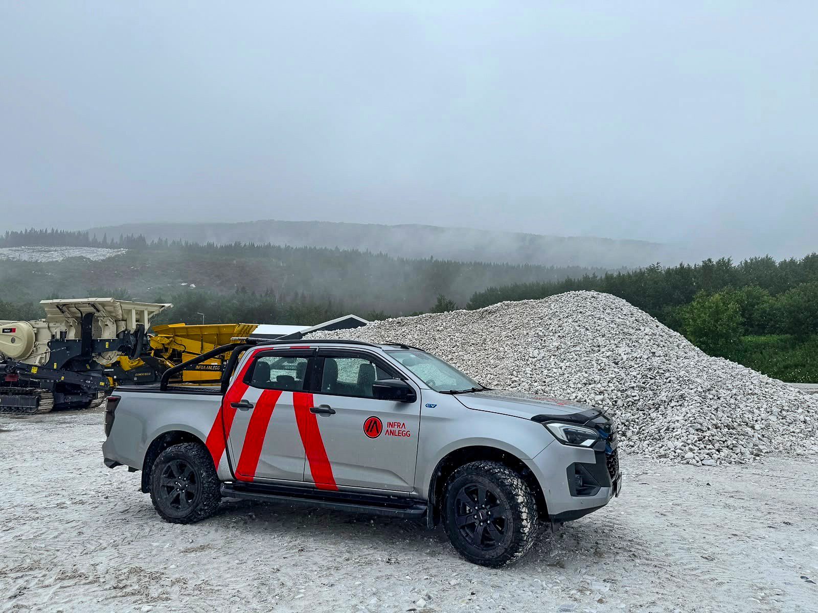 Silver pickup truck with red stripes and construction equipment next to a gravel pile and misty forest.